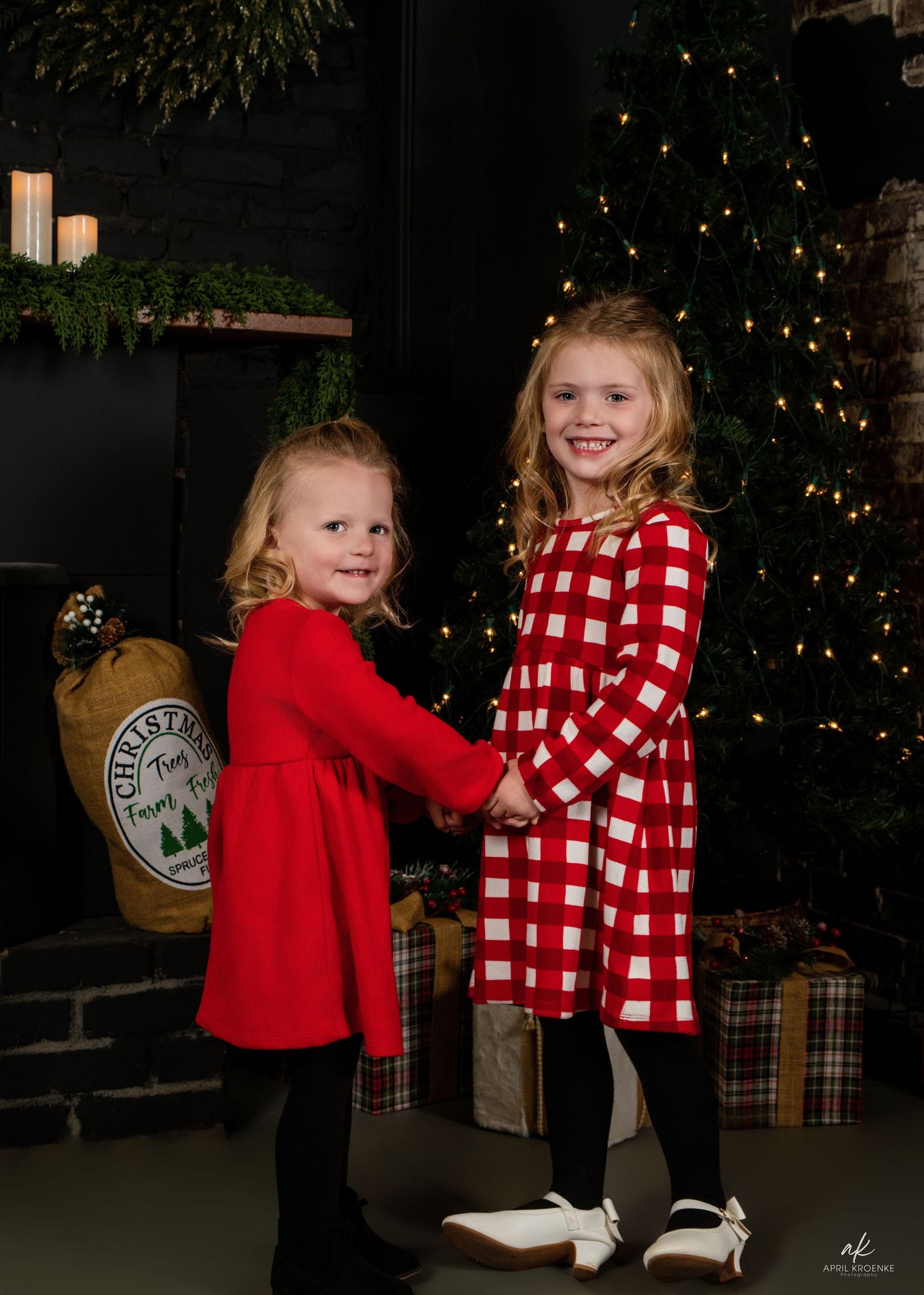 Two children in holiday attire pose together near a decorated Christmas tree in a dimly lit room.