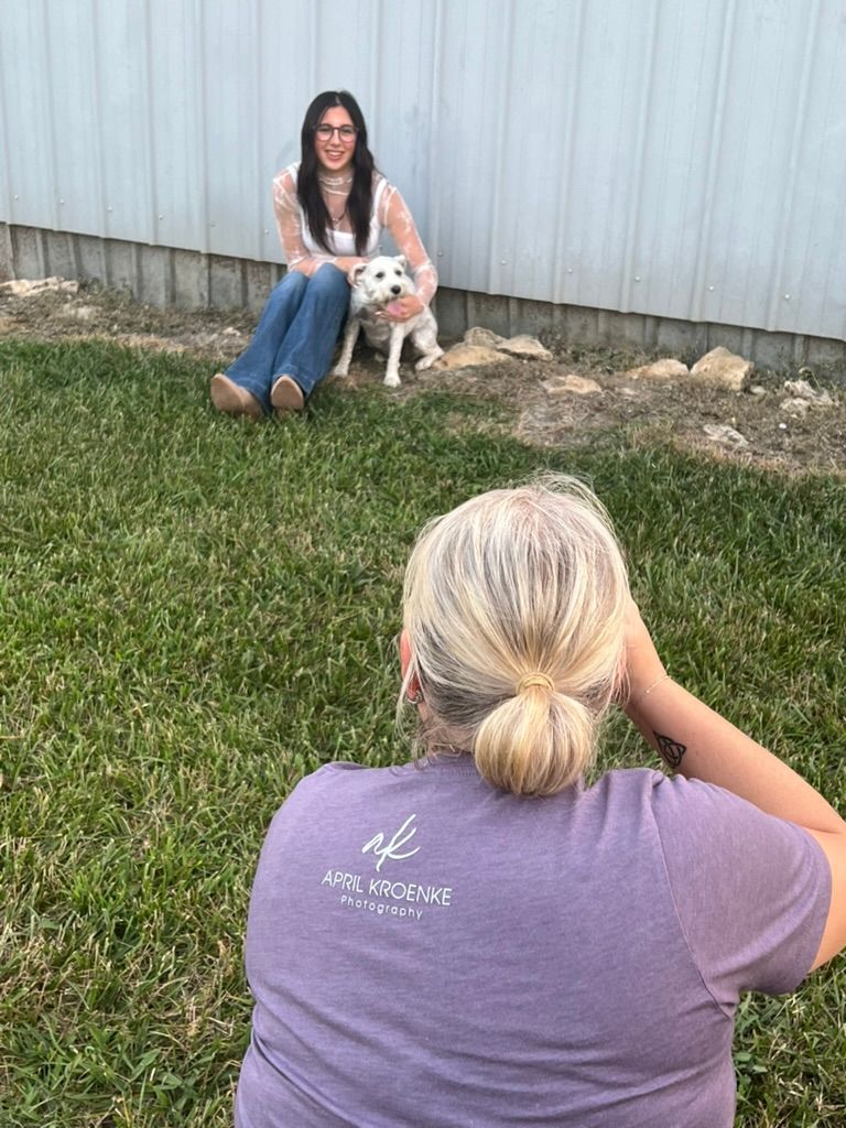 People sitting outdoors on grass with a small white dog near a metal fence.