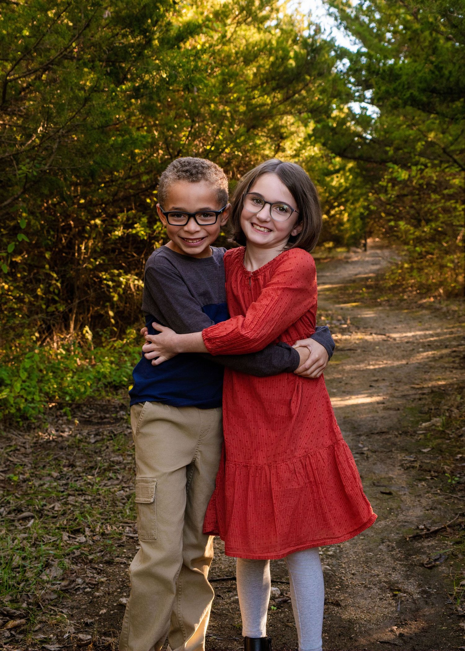 Two children embrace while standing on a forest trail with autumn foliage in background.