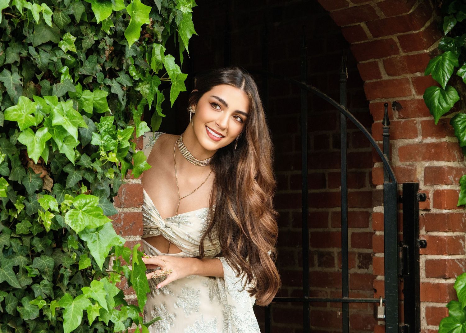 A bride in a white wedding dress stands next to ivy-covered brick wall, smiling at the camera.