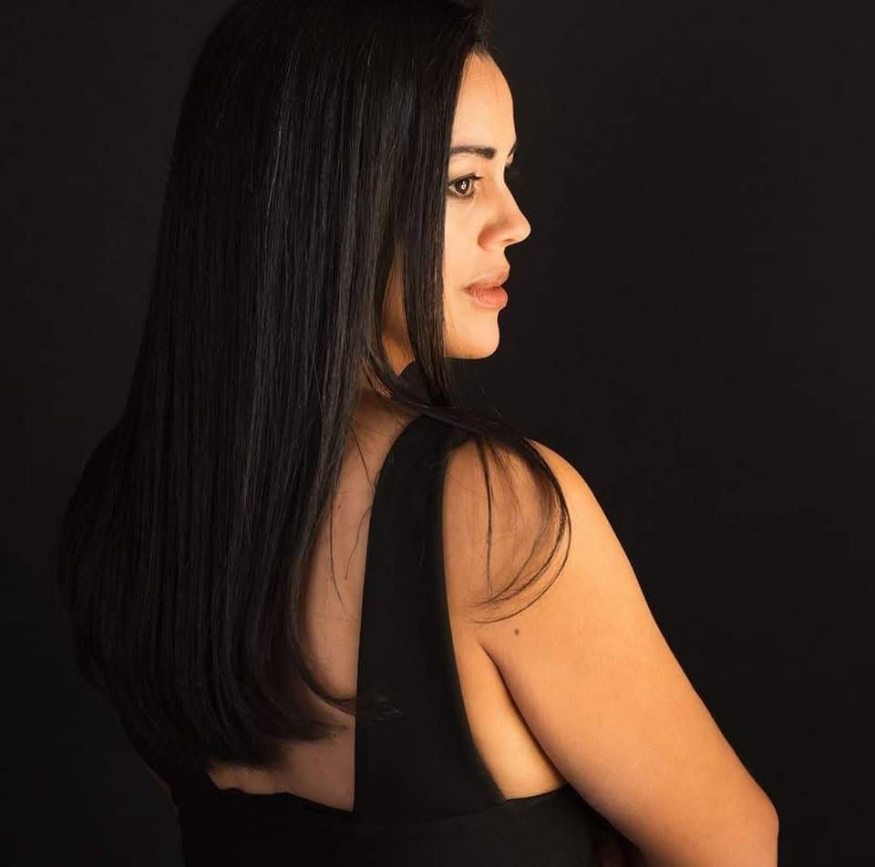 Woman with sleek black hair in profile view wearing a black backless top against a dark background.