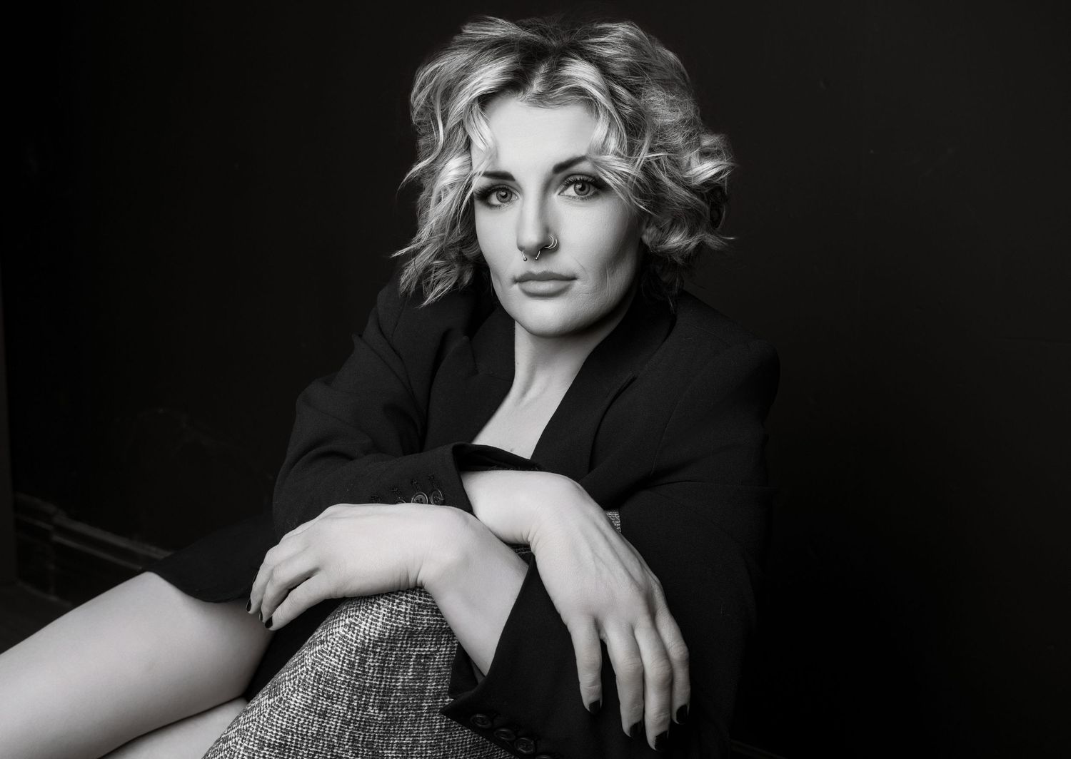Black and white portrait of a blonde woman in a black blazer sitting gracefully against a dark studio background.