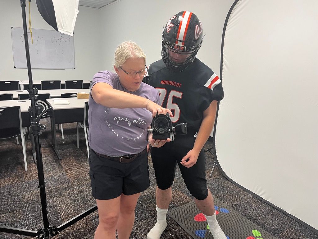A photographer adjusts lighting and camera settings while photographing a football player in uniform in a studio setting.
