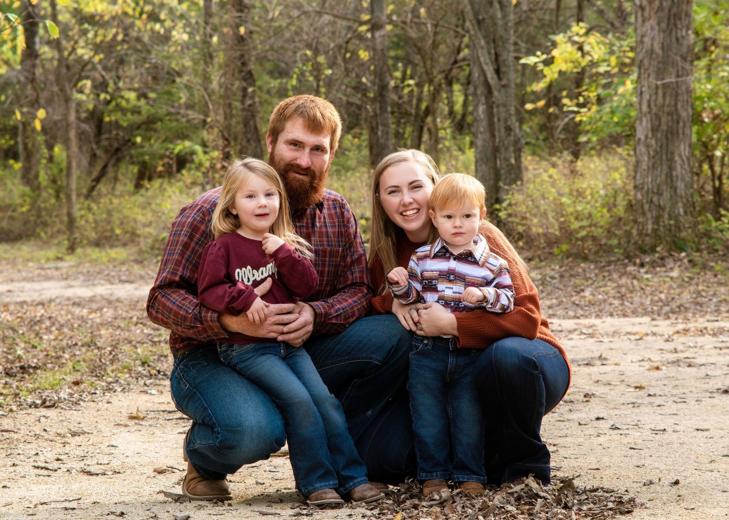 Family of four sitting together on dirt path surrounded by autumn trees.