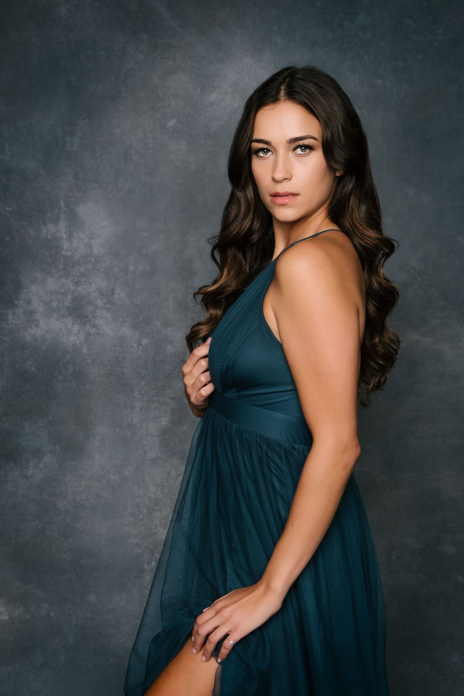 Young woman in teal halter gown poses elegantly against a dark gray studio backdrop with flowing brunette hair.