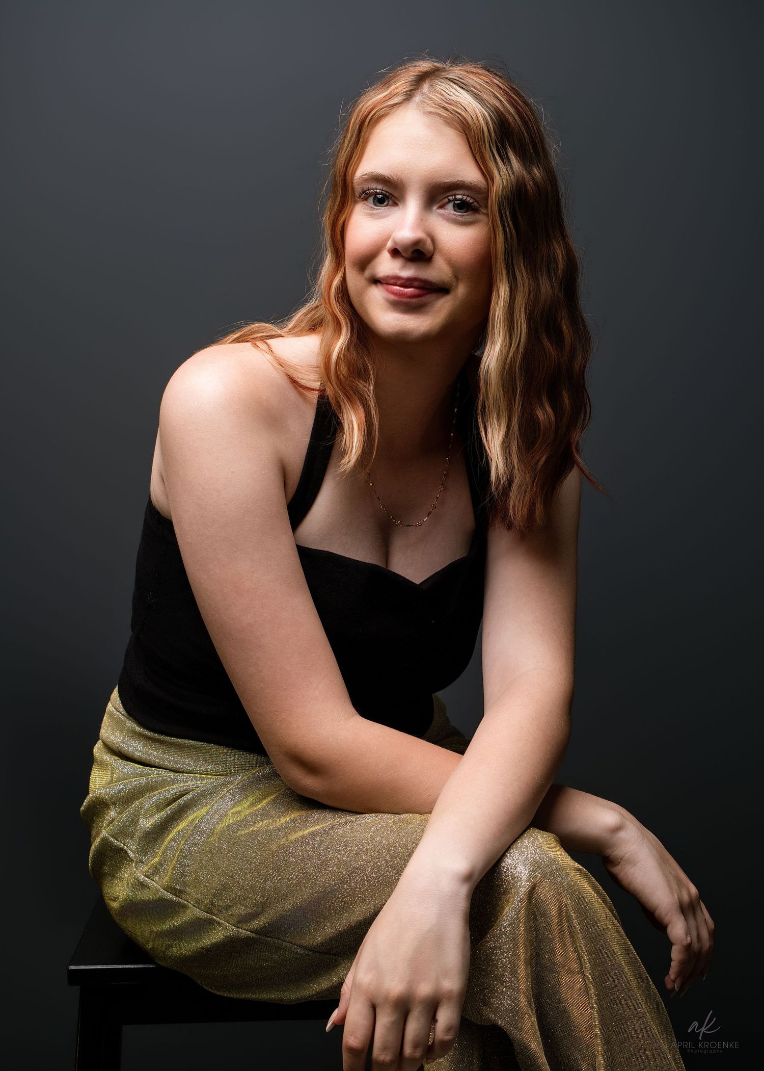 Young woman with auburn hair poses in black top and gold skirt, smiling confidently in a professional studio portrait.