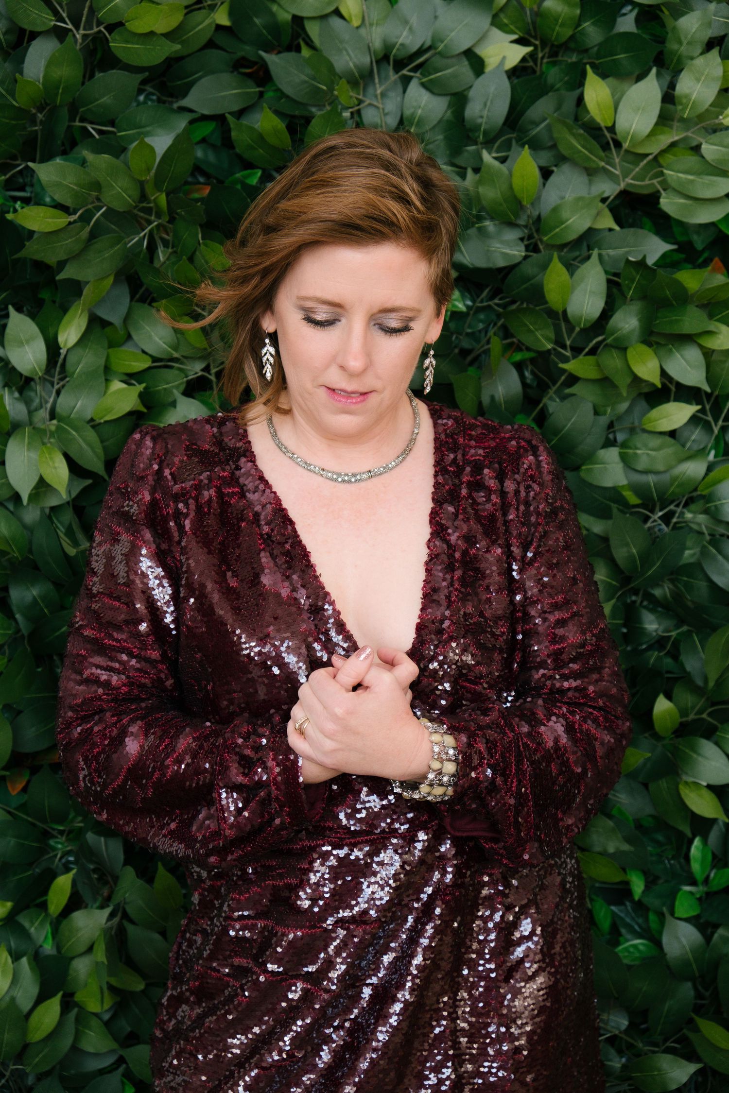 Woman in burgundy sequin dress with jewelry poses against lush green hedge backdrop.