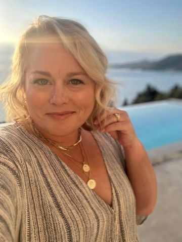 A person in a silver sequined top poses against a scenic coastal backdrop with an infinity pool and ocean view at sunset.