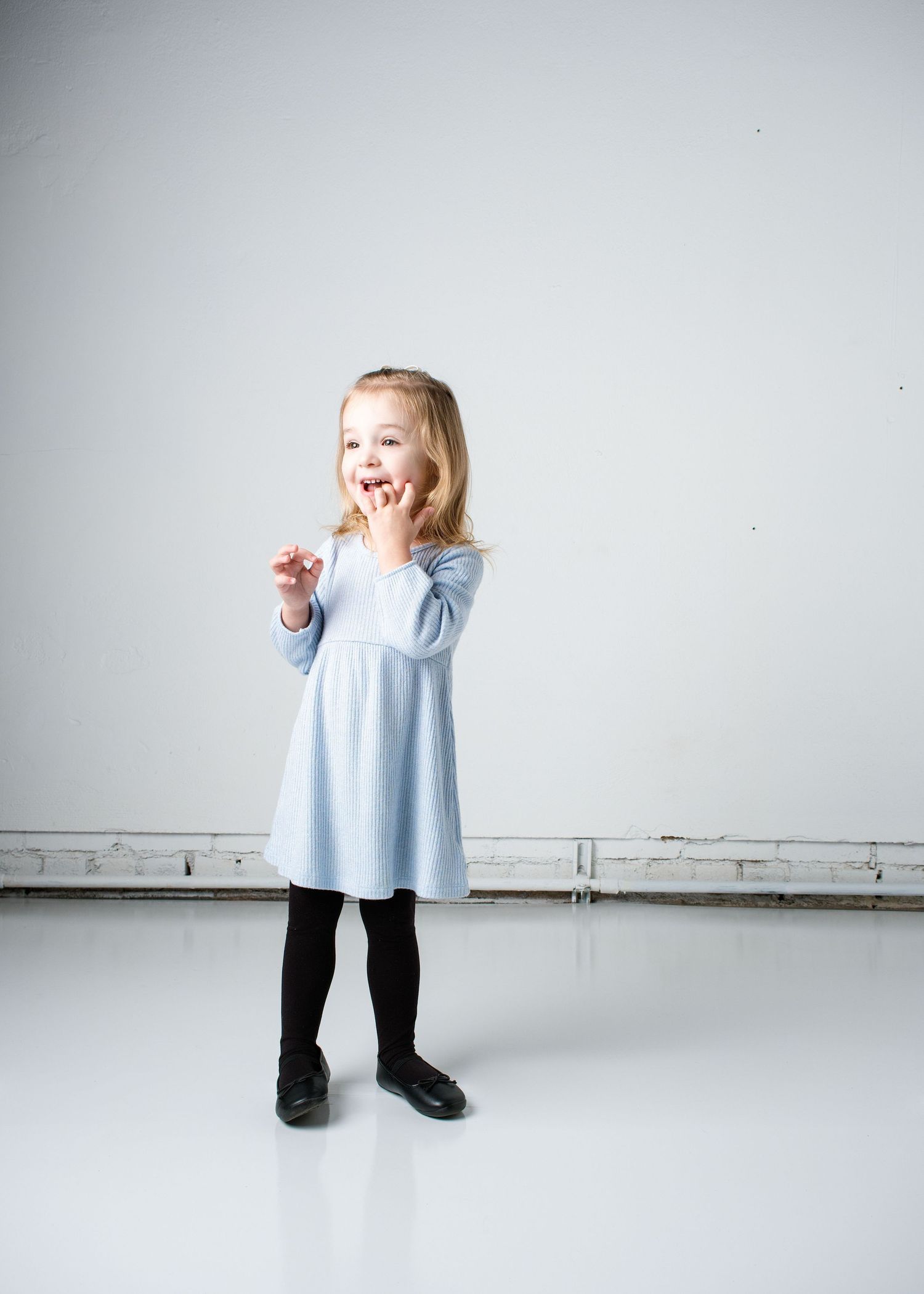Child in light blue top and black pants dances joyfully in minimalist white studio space.