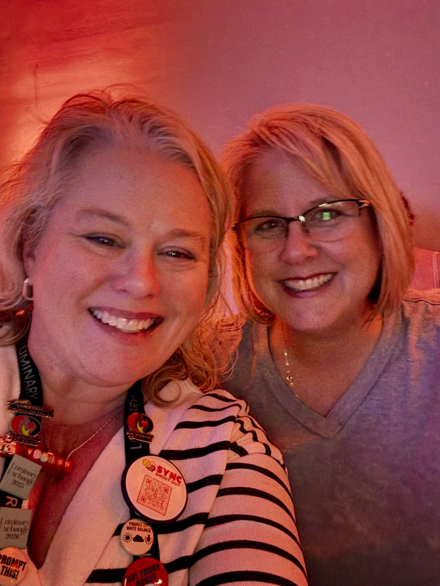Two people wearing glasses smile together in warm reddish lighting, one in a striped shirt with decorative pins.