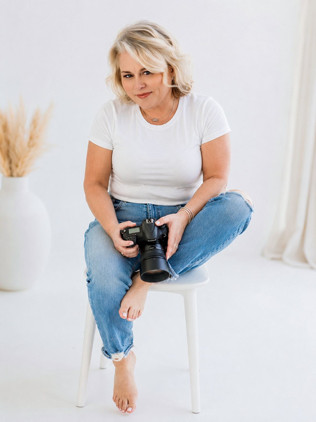 A blonde woman sits barefoot on a white stool holding a DSLR camera, wearing jeans and a white t-shirt.