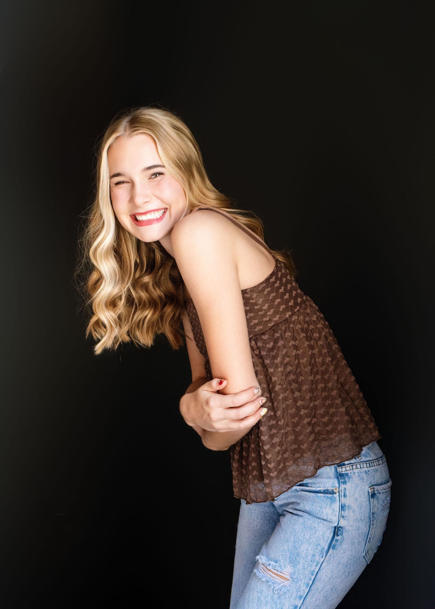 A person wearing a brown knit sweater and blue jeans poses playfully against a dark background.