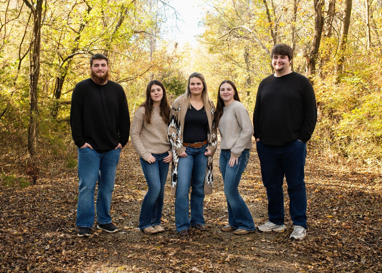 Group of five people in casual wear pose together on leaf-covered path in autumn woods.