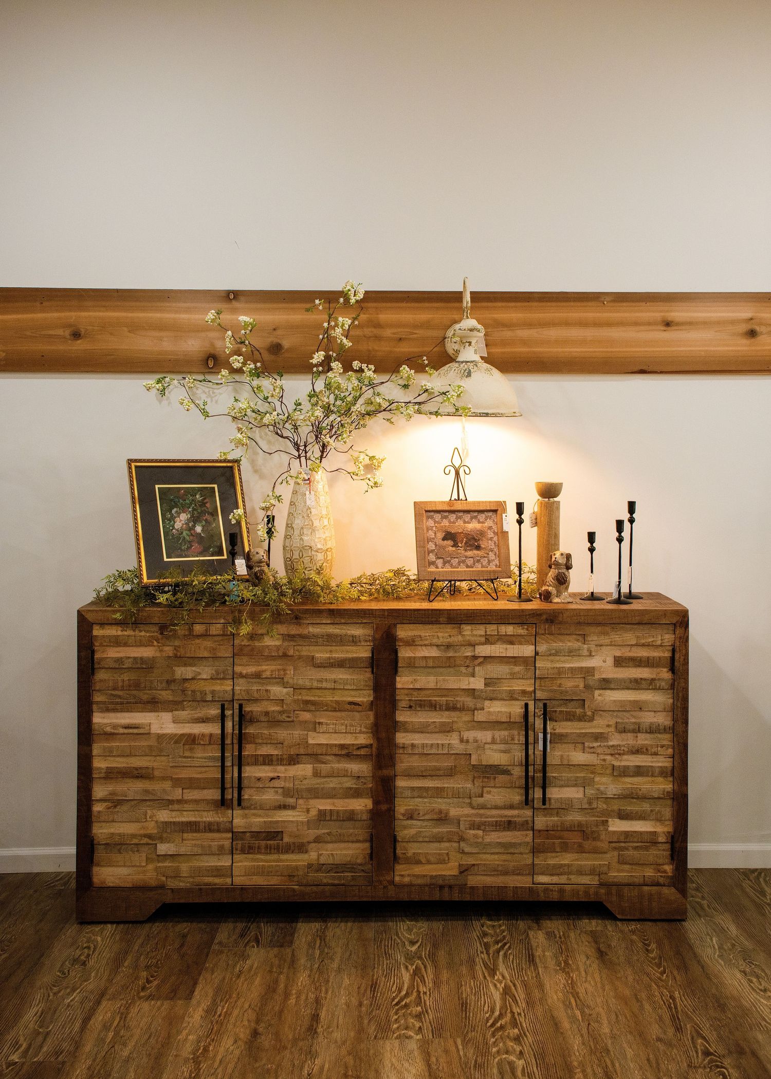 Rustic wooden sideboard with textured panels styled with table lamp, candlesticks and greenery against neutral wall.