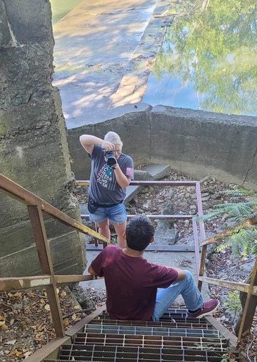 Two people sit on wooden steps leading down to a creek or stream while enjoying a peaceful outdoor moment.