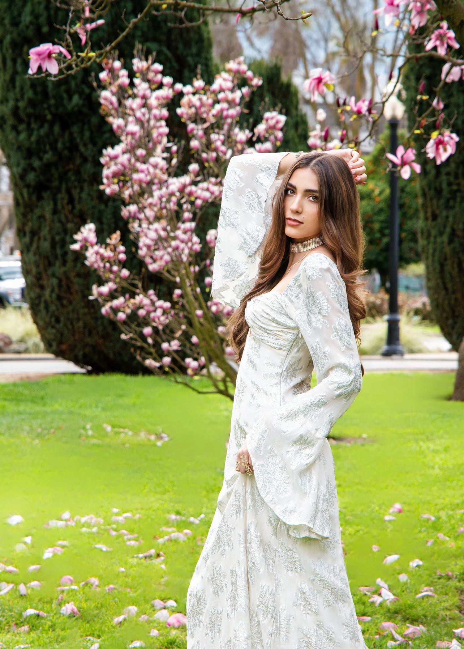 A figure in a flowing white lace dress poses gracefully among blooming pink magnolia trees in a spring garden setting.
