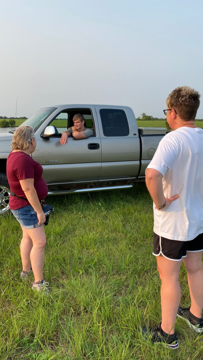 Several people examine something in tall grass next to a parked silver pickup truck on a sunny day.