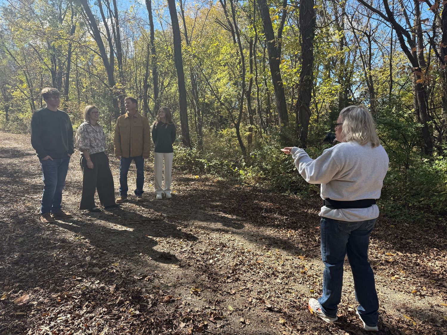 People walking along a wooded nature trail on a sunny autumn day with fallen leaves covering the ground.