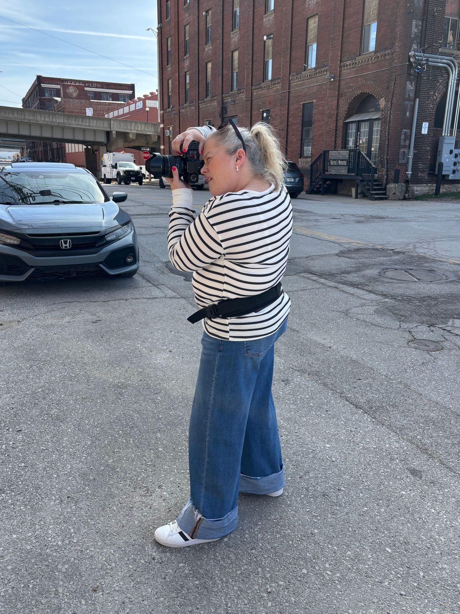 Person in striped shirt and jeans takes photos in an industrial parking lot with brick buildings in the background.