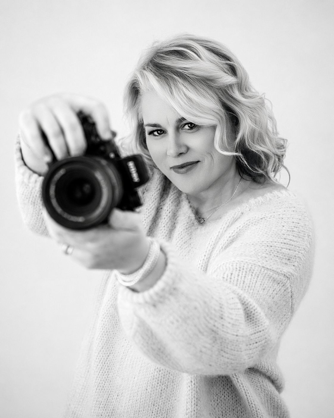 Black and white portrait of a blonde woman holding a Nikon camera toward the viewer, wearing a cozy white sweater.