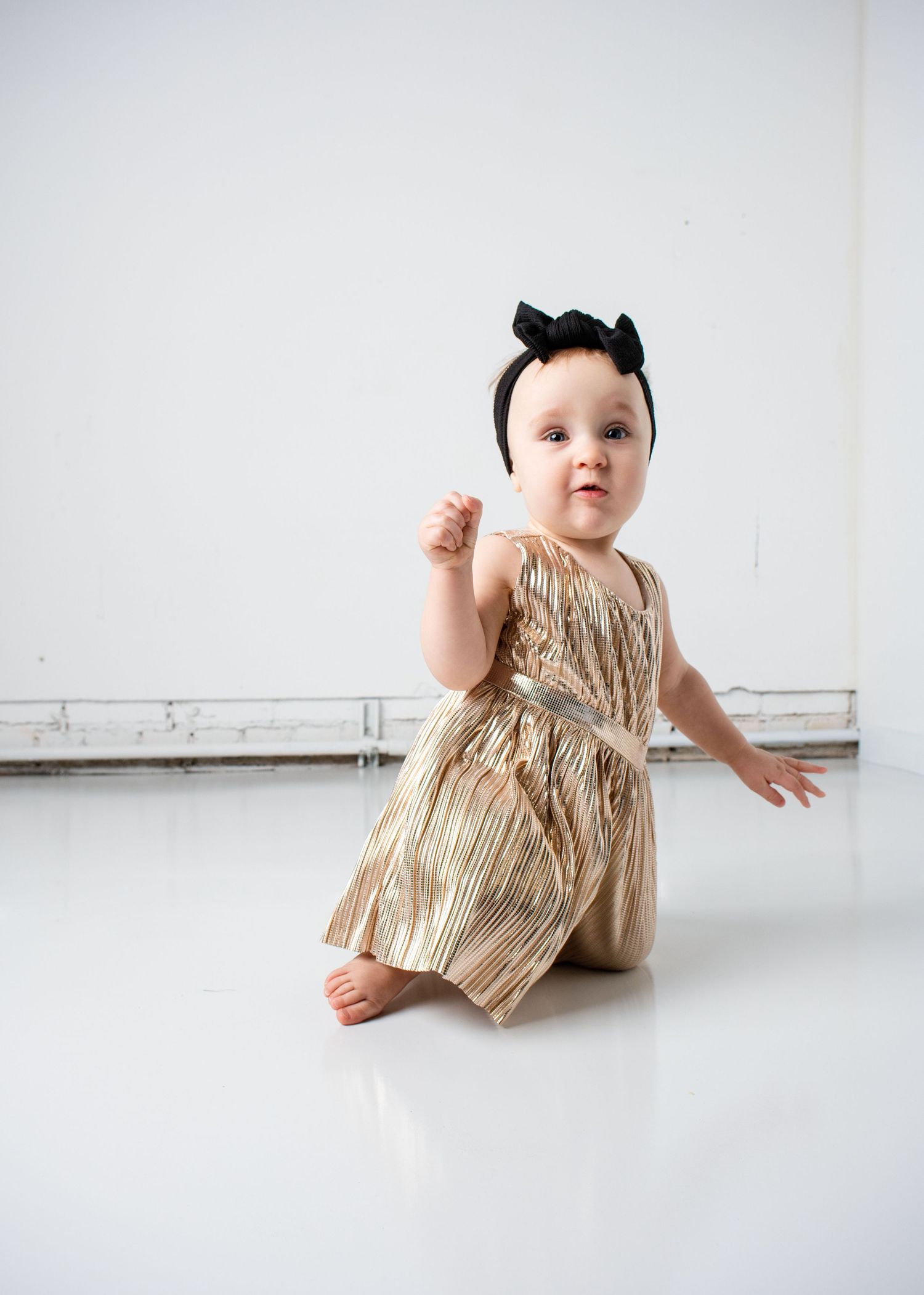 Baby in gold dress and black bow sits on white floor during studio photoshoot.