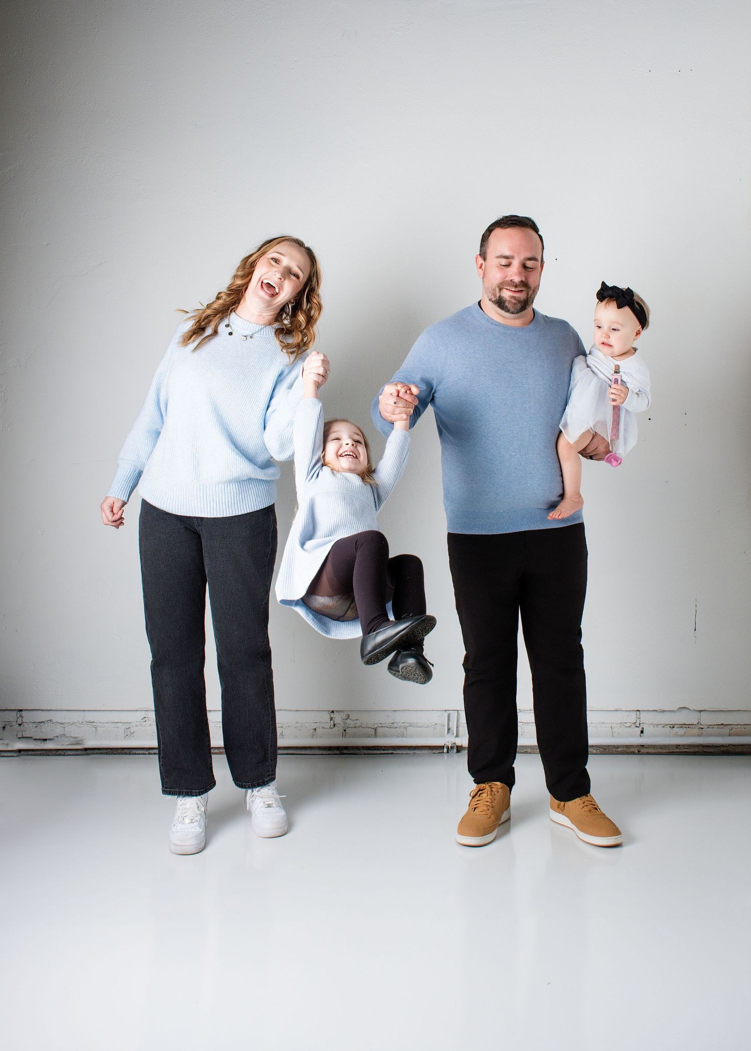 Family of four wearing casual clothes jumping and posing playfully against a white wall in a studio setting.