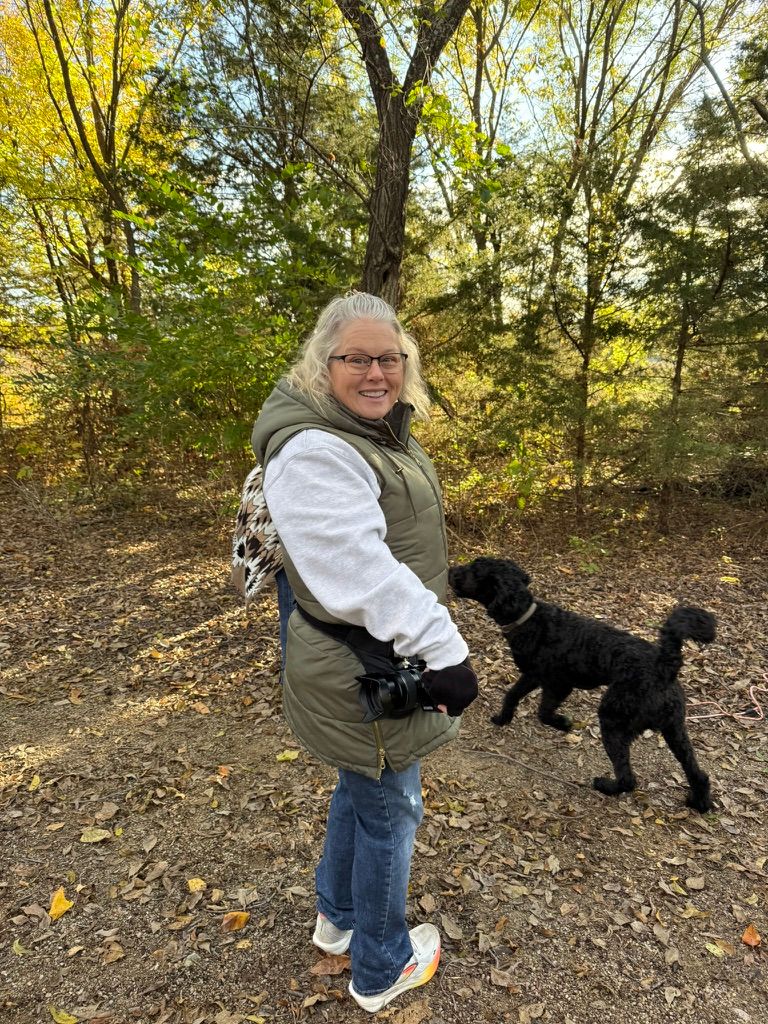 A black dog plays in autumn leaves on a wooded trail while someone watches from nearby.