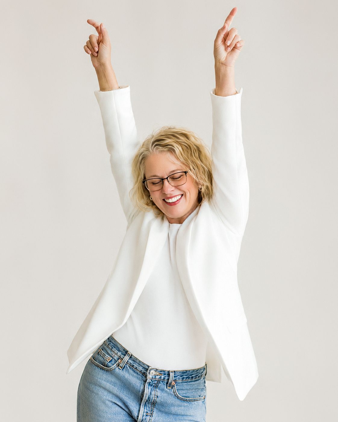 A cheerful blonde woman in a white blazer and jeans raises her arms in celebration against a light background.