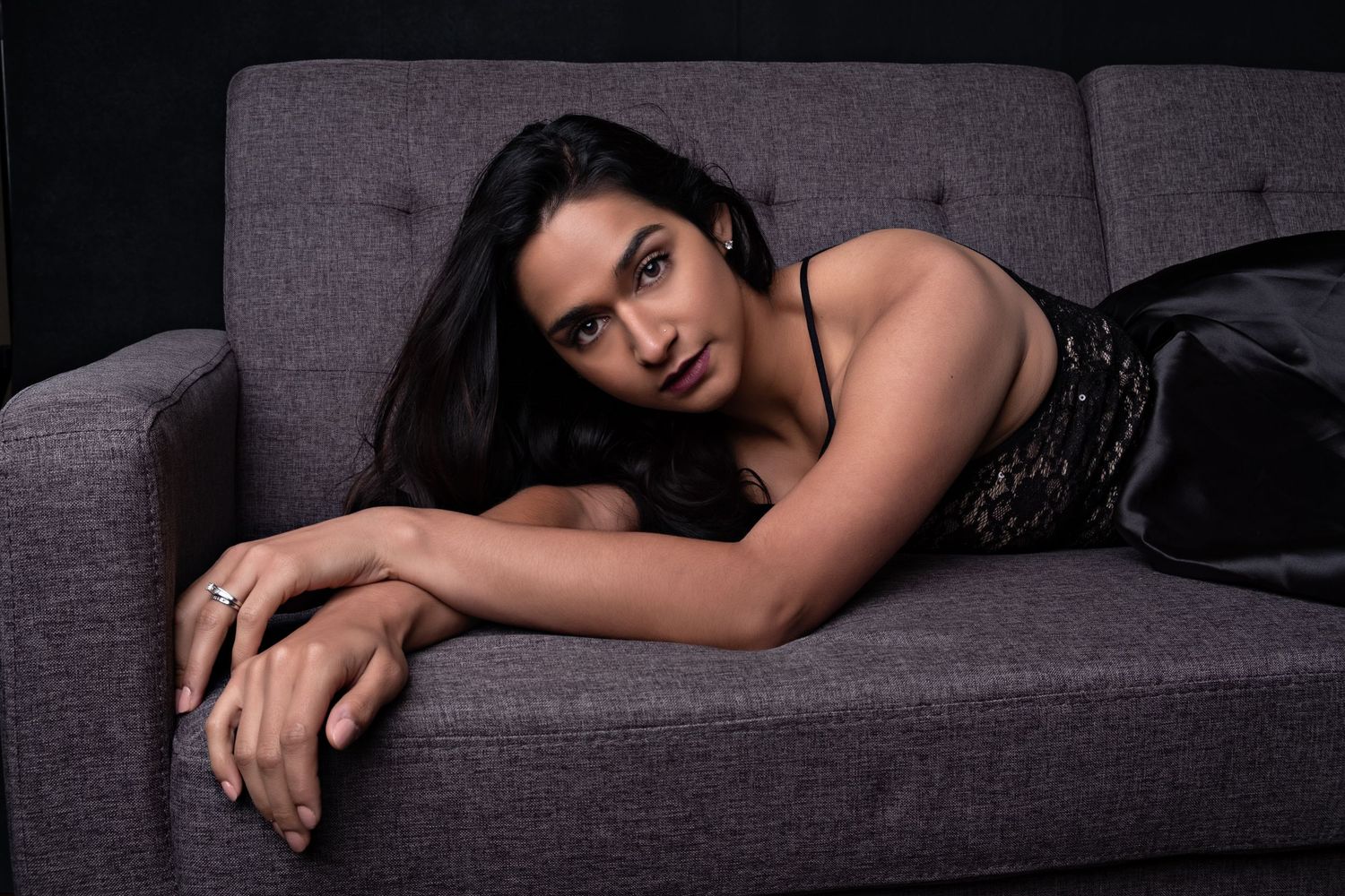 A woman in a black dress lying on a grey sofa, gazing at the camera in a moody studio setting.