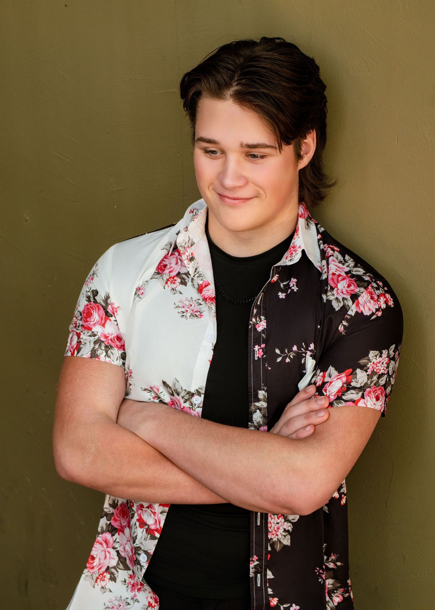 Young man in floral shirt with arms crossed, smiling against green wall.