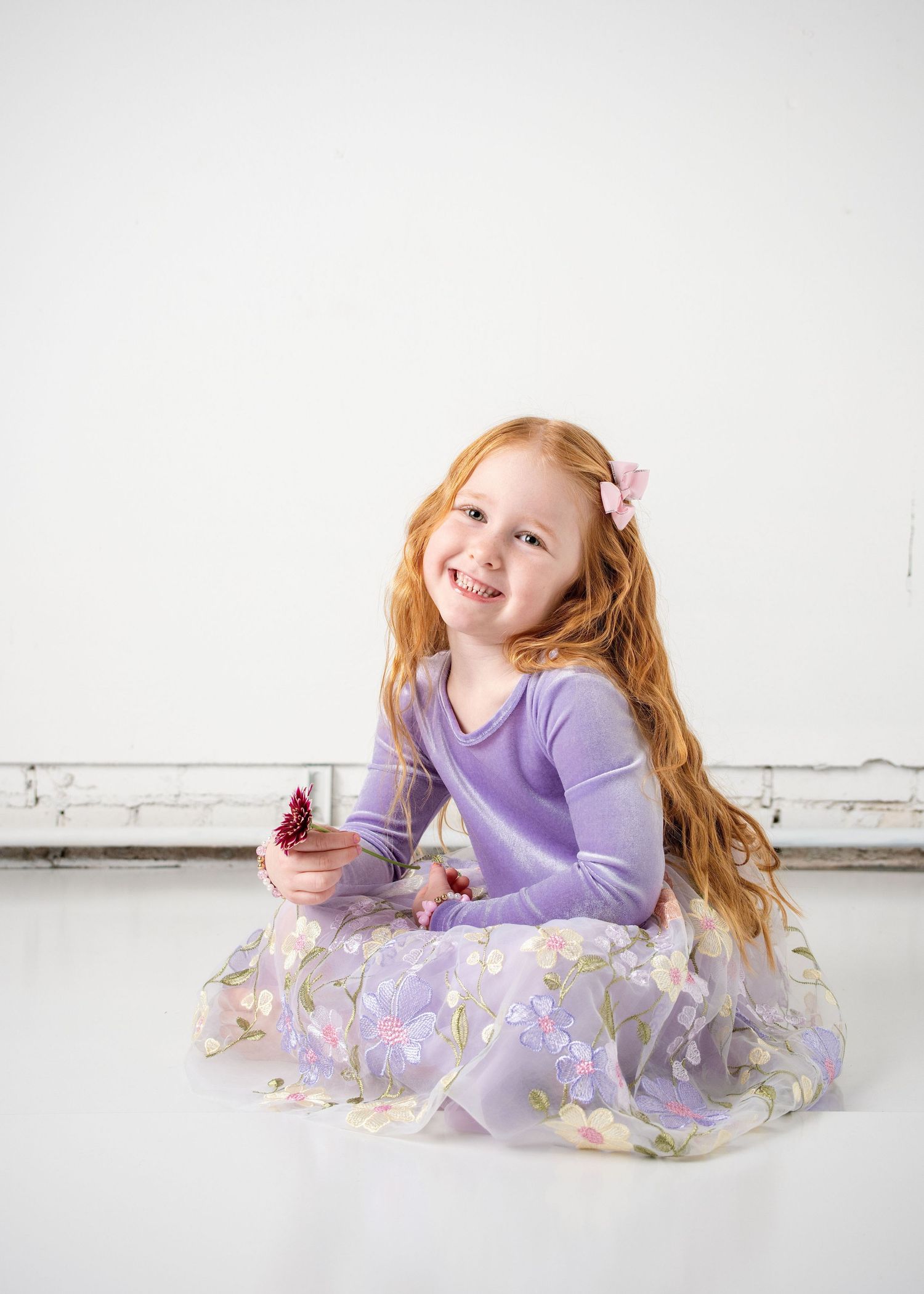 Young dancer in purple leotard and floral tutu performs graceful floor poses against white background.