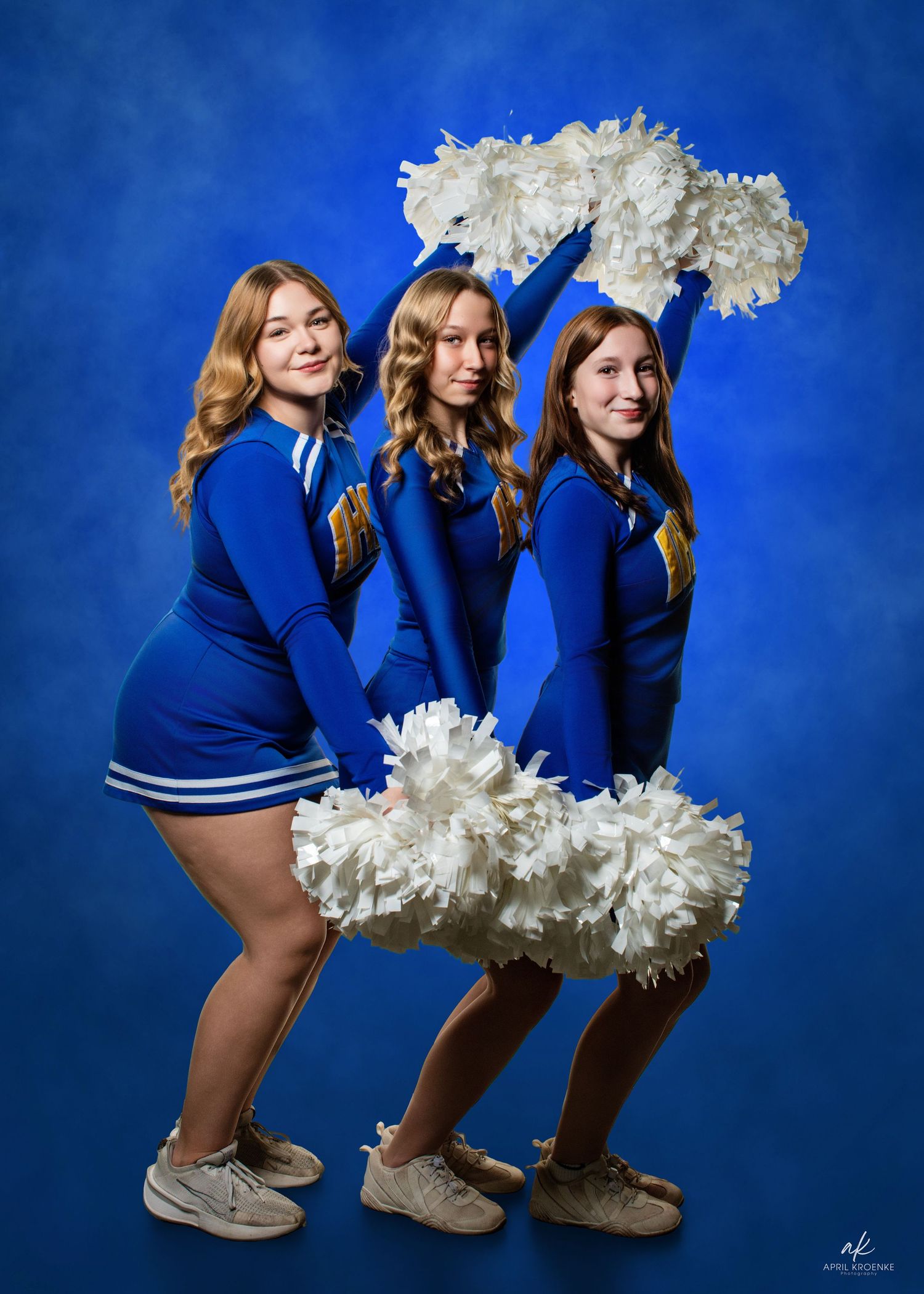 Cheerleaders in blue uniforms with white pom poms perform synchronized routine against blue backdrop.