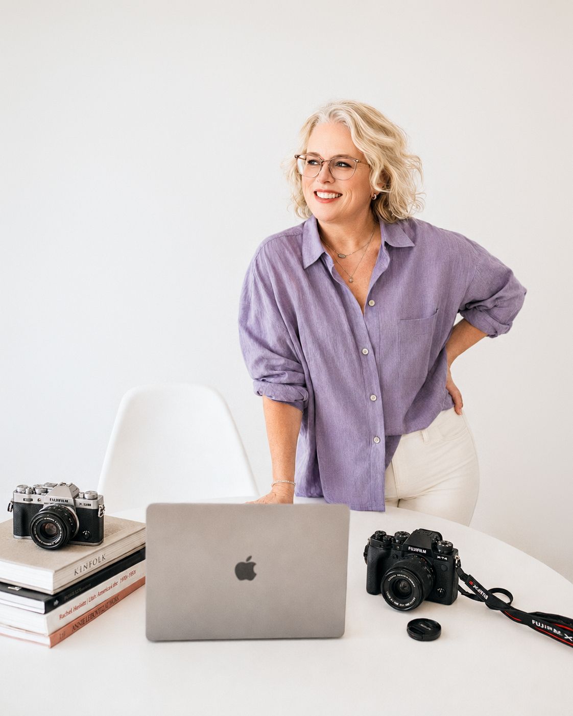 Woman in purple shirt smiling beside a MacBook and cameras on a white desk in a bright studio.