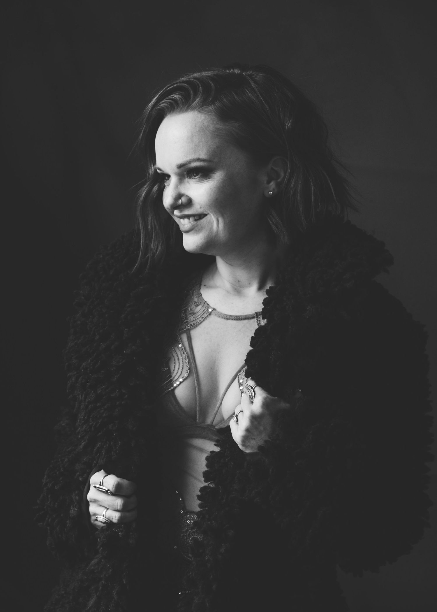 Black and white portrait of a smiling woman in a fluffy coat, looking sideways in a dark studio setting.
