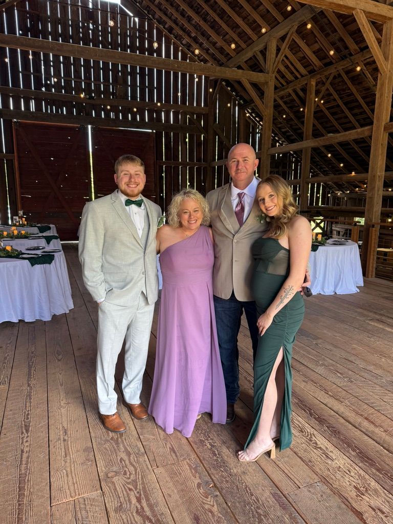 Wedding party members in formal attire stand together inside a rustic wooden barn venue with natural lighting.