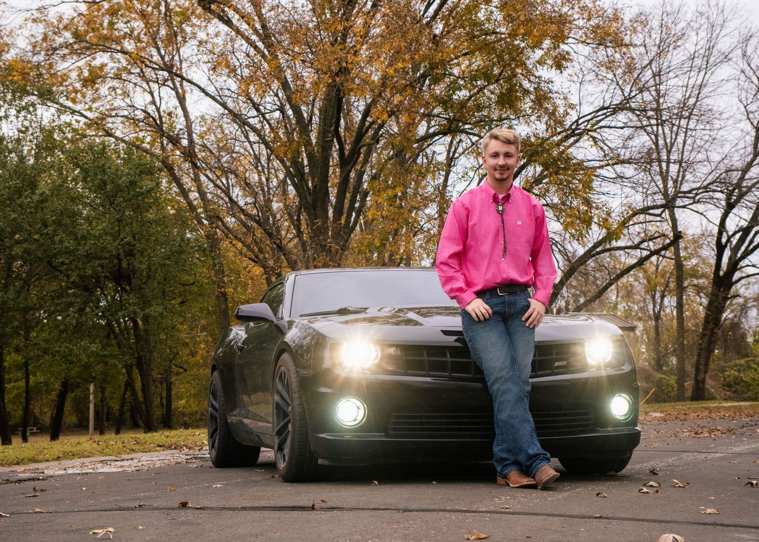 Black car with headlights on parked on autumn road with fallen leaves at sunset.