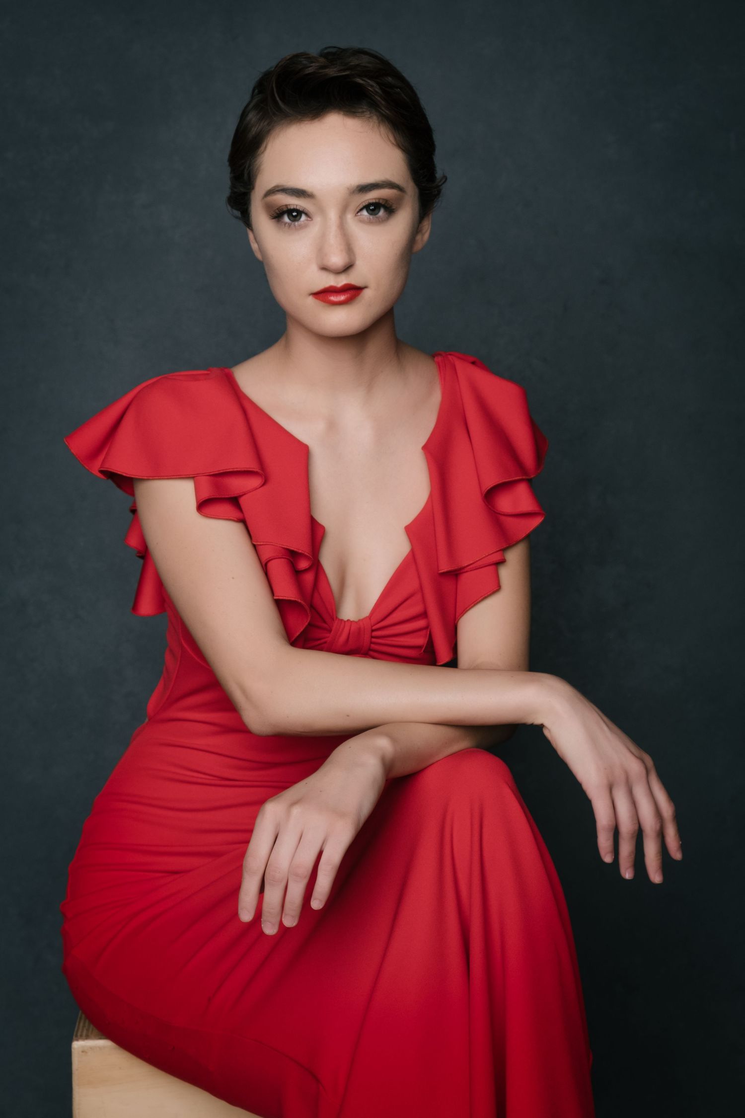 Young woman in elegant red ruffled dress with deep V-neckline, seated gracefully against dark background.
