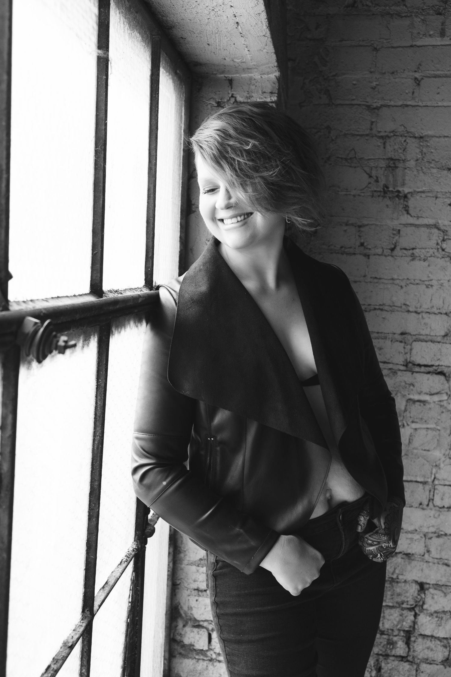 Smiling woman in black jacket leaning against brick wall by window in black and white portrait photo.