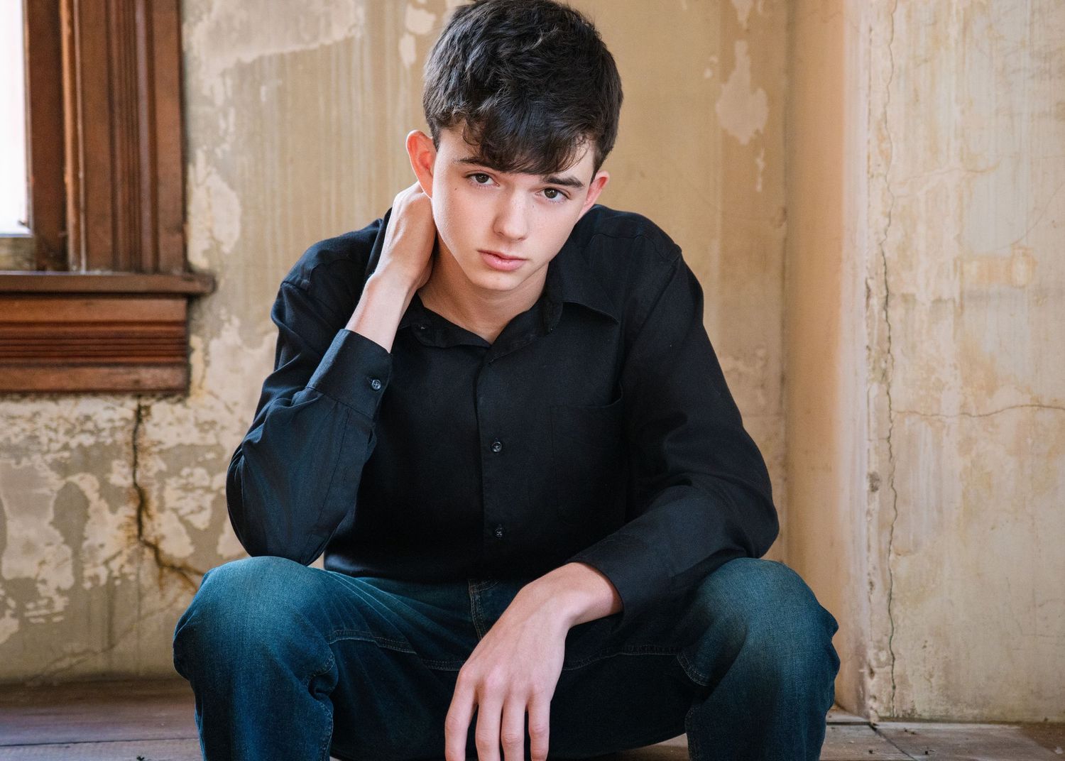 A young man in a black shirt and jeans sits against a rustic wall, resting his head on his hand.