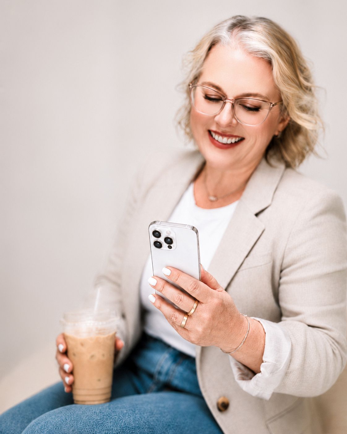 Smiling woman in beige blazer using smartphone while holding iced coffee drink.