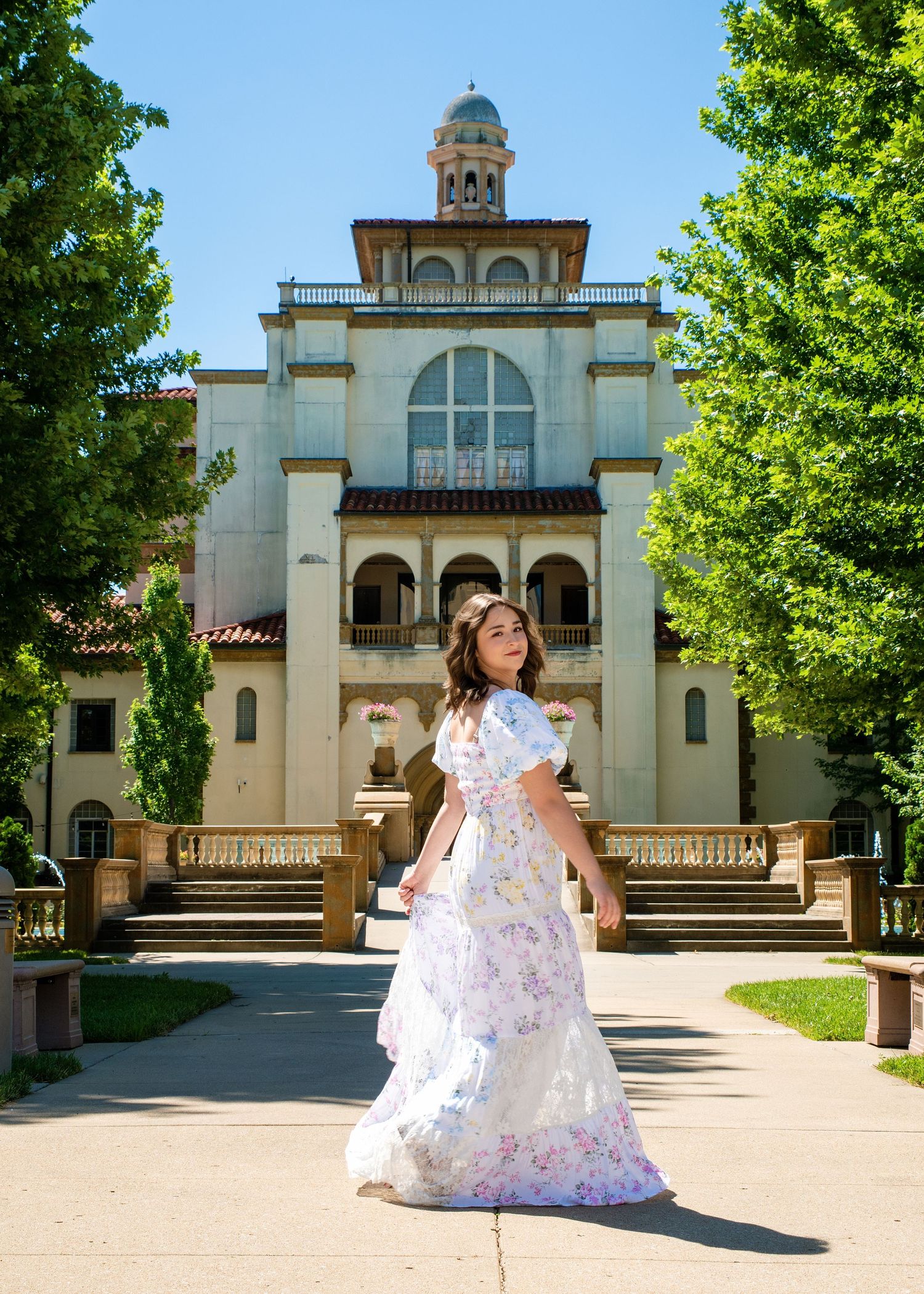 Young woman in floral white gown poses on pathway before elegant historic building with dome and lush green trees.