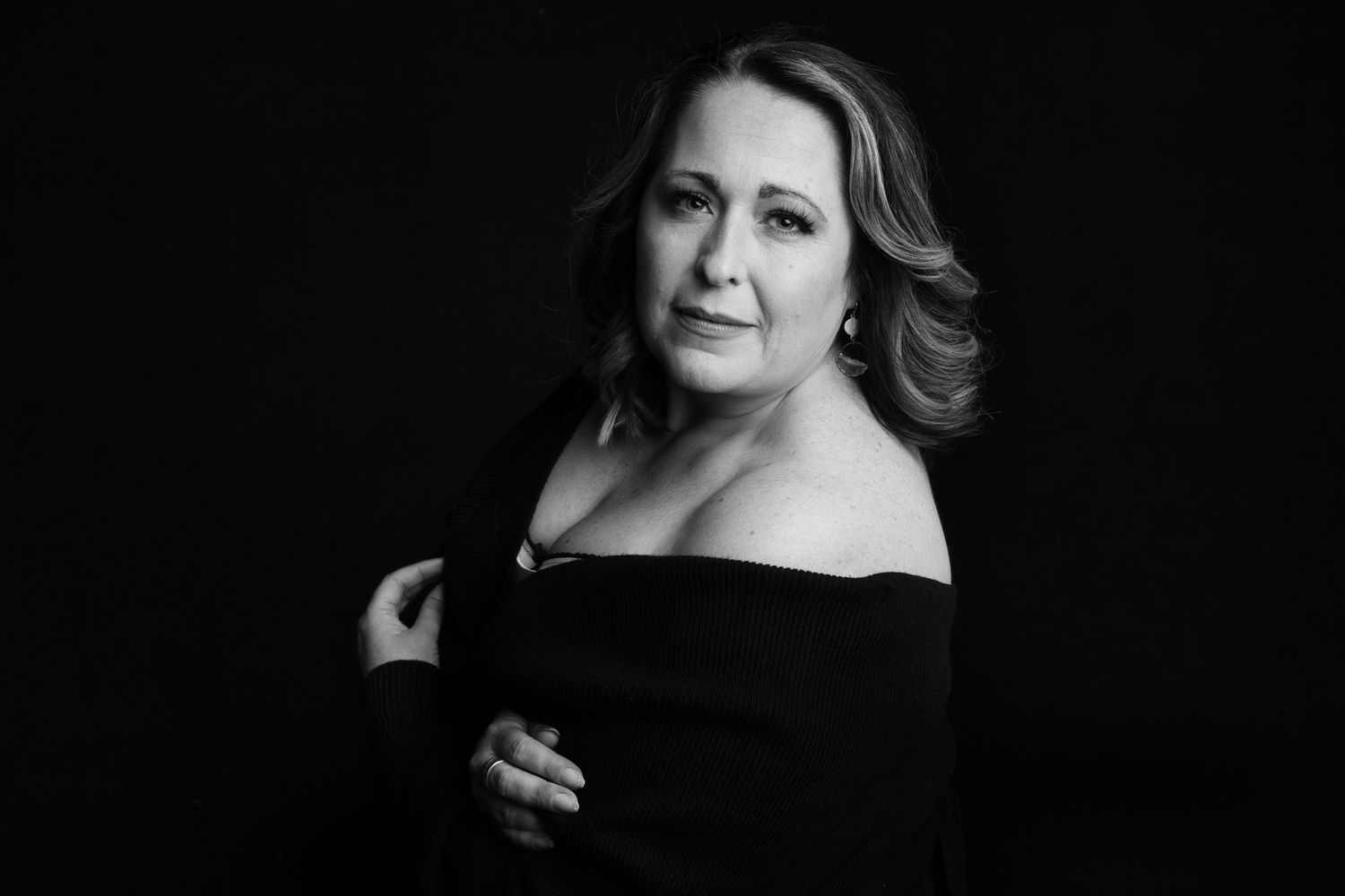 Black and white portrait of a woman in an off-shoulder black top, posing elegantly against a dark background.