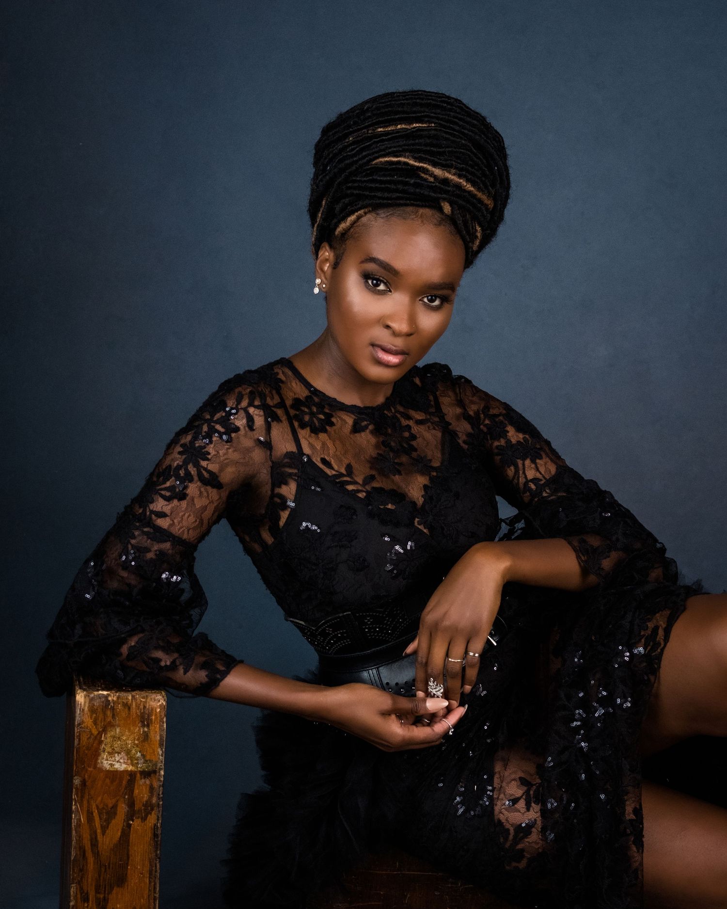 A woman in black lace dress and gold-trimmed head wrap poses elegantly against a dark blue studio background.