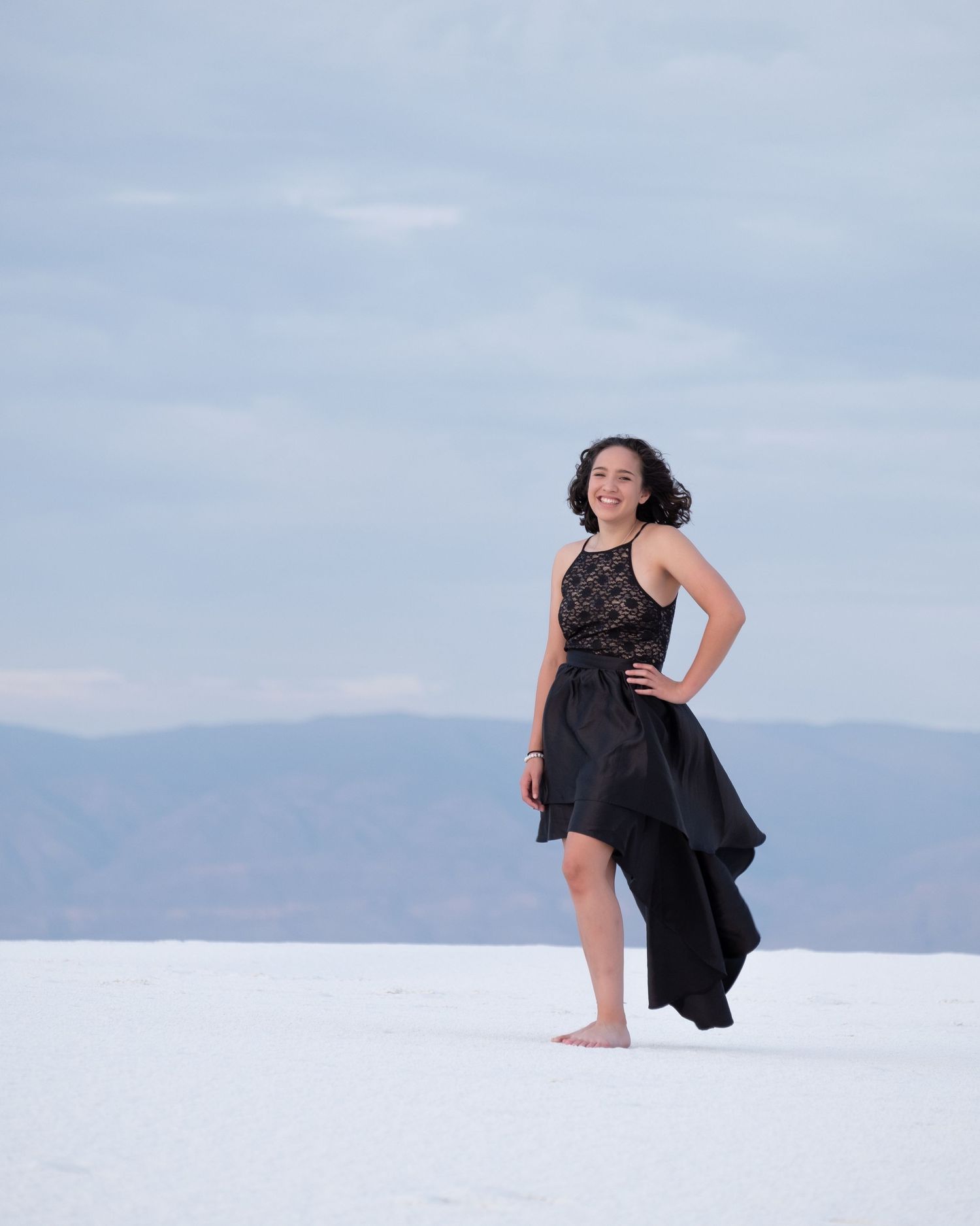 A person in a black dress poses gracefully against a stark white sand landscape with mountains in the distance.