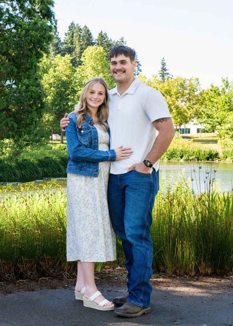 A couple stands together by a scenic pond on a sunny day, with lush greenery and trees in the background.