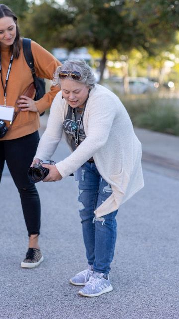 A person in a white sweater and jeans bends down to tie their shoe on a paved path.