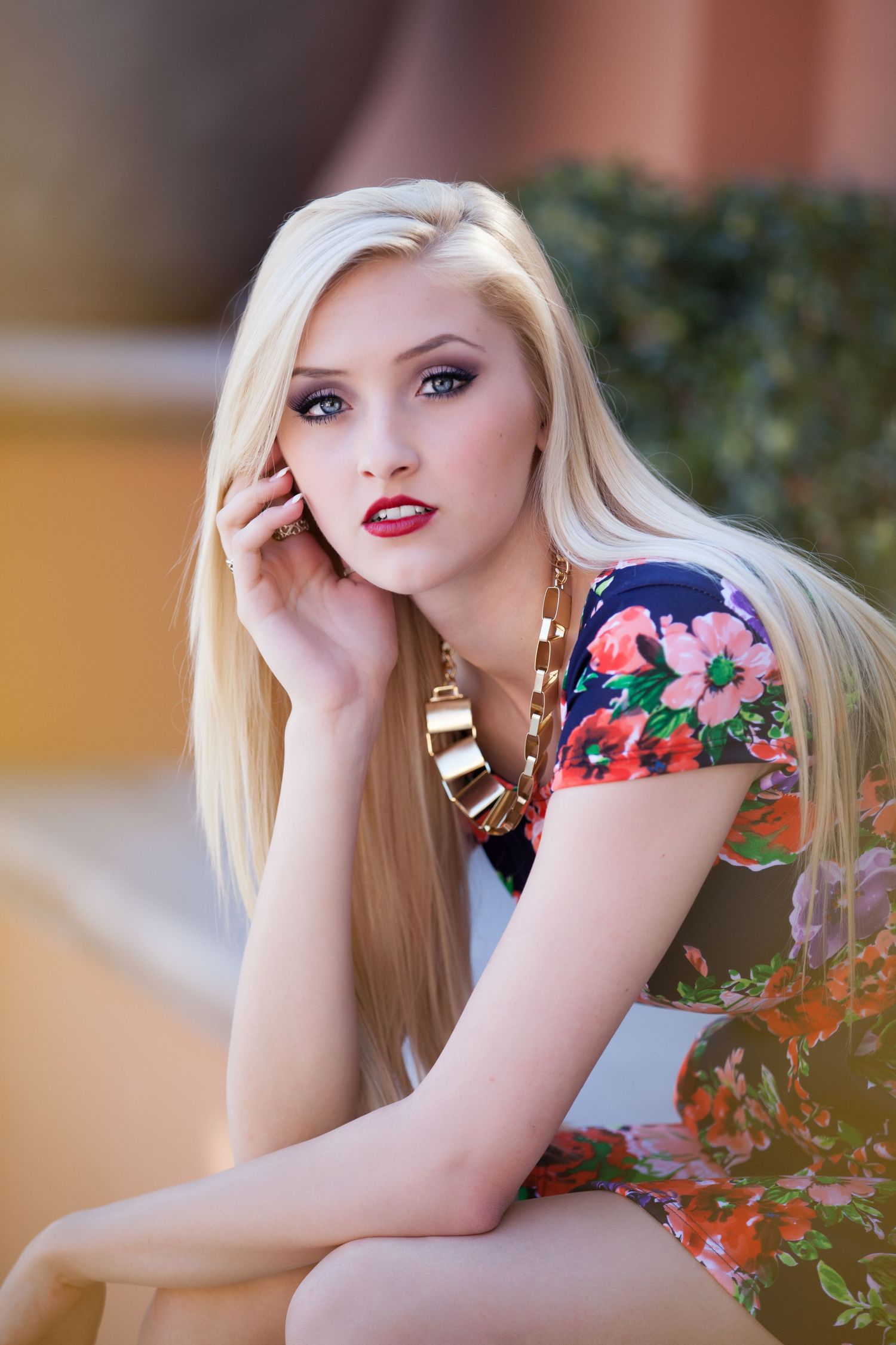Portrait of someone with long blonde hair wearing a floral dress and gold statement necklace in an outdoor setting.