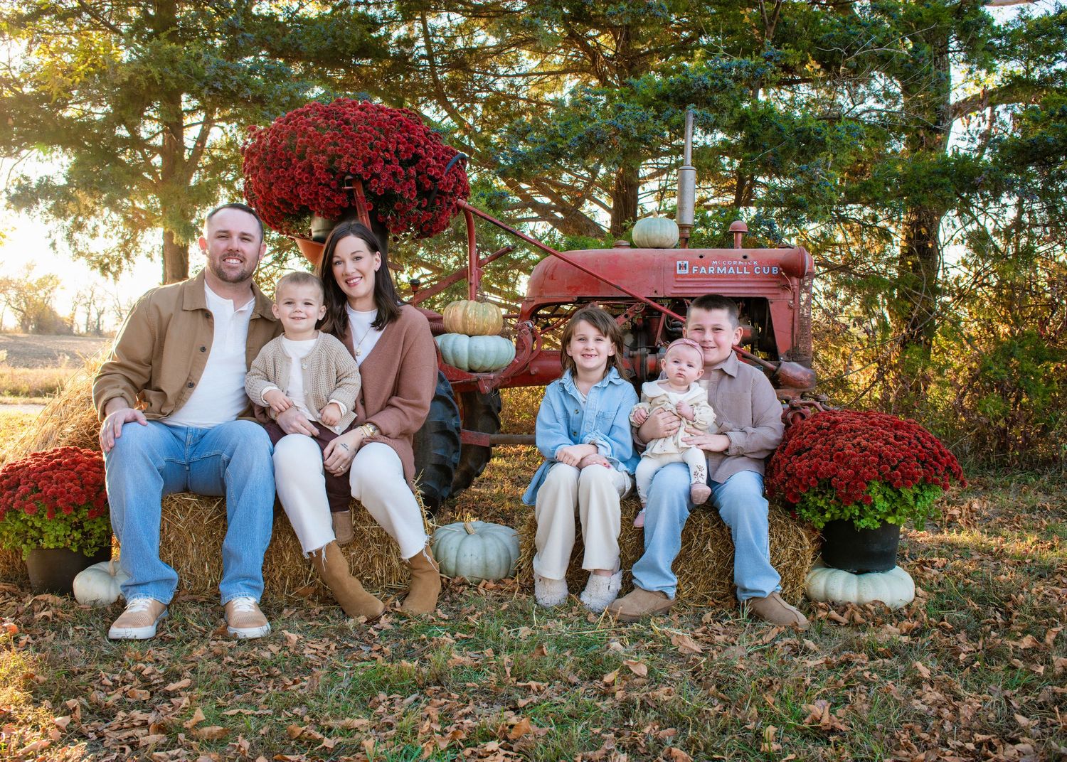 Family members sitting together on fallen autumn leaves with red bushes and trees in the background.