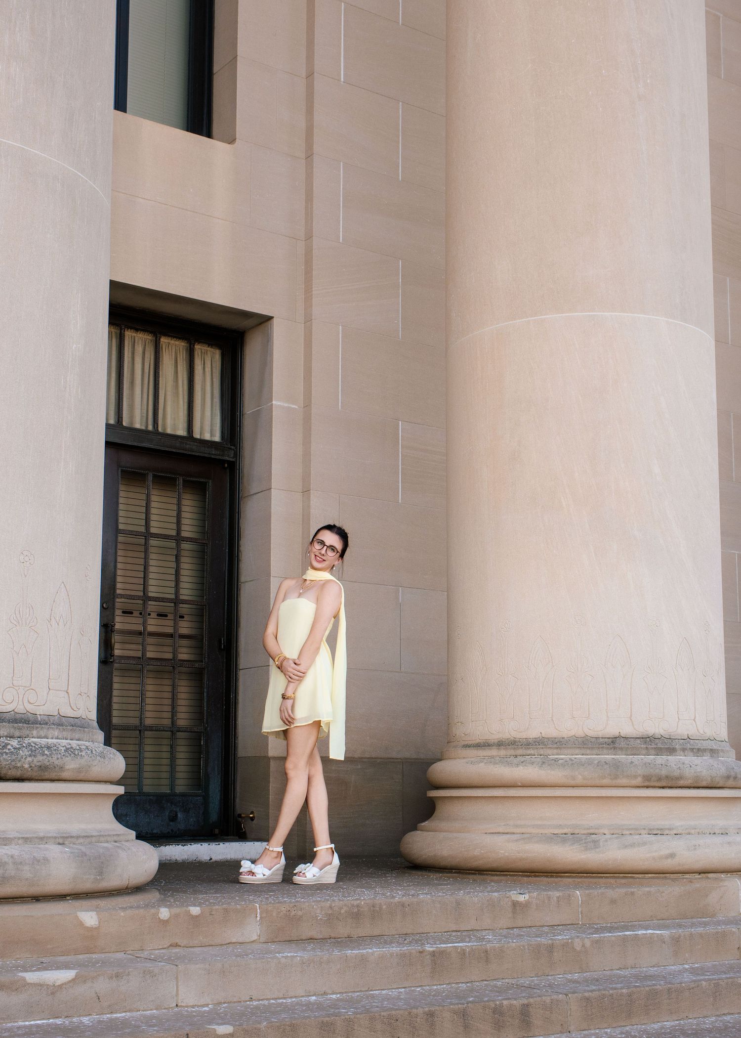 Woman in yellow dress posing by large stone columns of a classical building on sunny day.