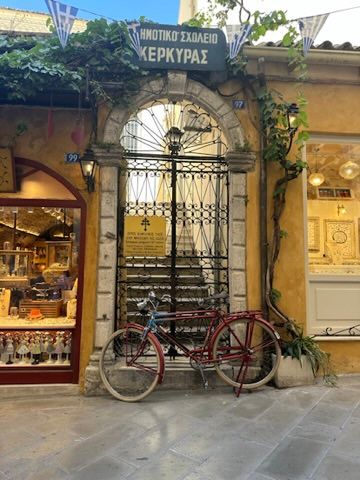 A vintage red bicycle parked outside a traditional Greek storefront with yellow walls and ornate iron window details.
